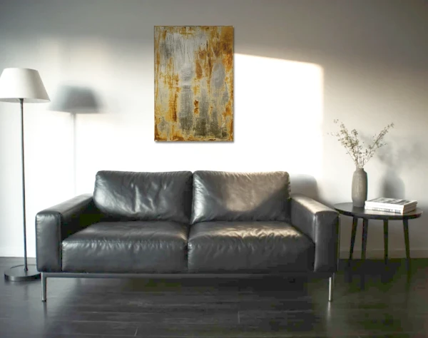 Eine Leinwand mit rostigen Flächen zwischen einer ansonsten silberglänzenden Lackfläche, hängt über einem schwarzen Ledersofa. – mixed-art
